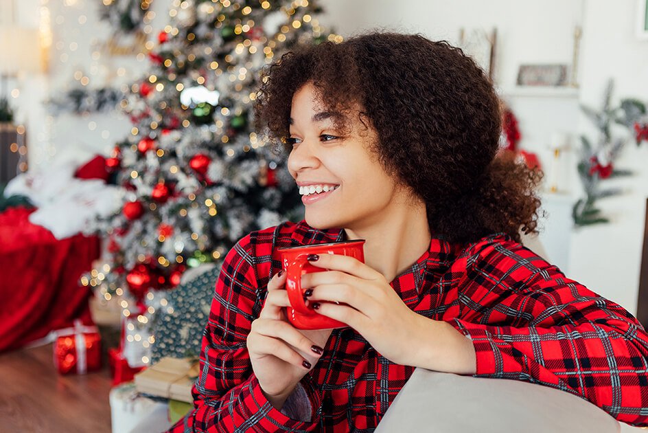 Woman holding up cup as she sits near a Christmas tree. 